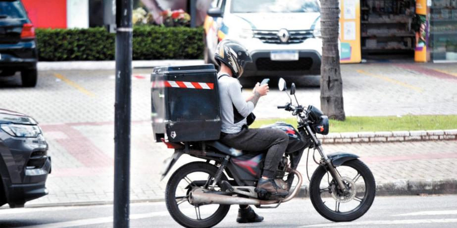 A imagem mostra uma pessoa pilotando uma motocicleta em uma rua. A motocicleta tem uma grande caixa acoplada na parte traseira, provavelmente para fins de entrega. O motociclista está usando um capacete e parece estar usando um smartphone com uma mão enquanto manobra a motocicleta com a outra. Há veículos ao fundo, indicando que esta cena ocorre em uma área urbana com tráfego. Esta imagem pode ser interessante ou relevante, pois captura um momento de multitarefa do motociclista, o que pode levantar preocupações sobre segurança no trânsito.