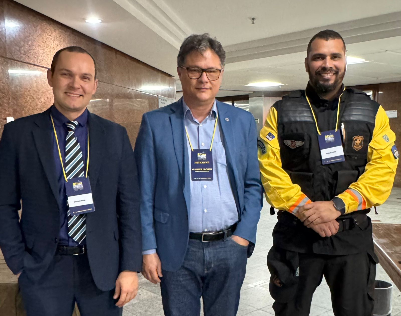 A imagem apresenta três homens lado a lado em um ambiente interno bem iluminado, com uma parede de mármore marrom polido ao fundo.