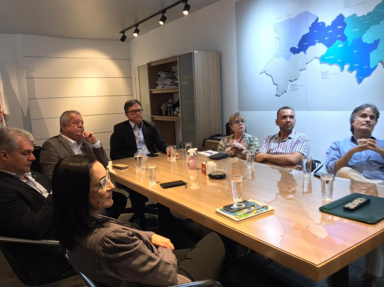A mesma reunião da primeira imagem, mas capturada de um ângulo levemente diferente, mostrando mais detalhes da mesa e das pessoas presentes. No fundo, há um mapa geográfico estilizado em azul na parede. A mesa contém copos de água e itens como cadernos e smartphones. Novamente, os participantes estão focados na reunião.