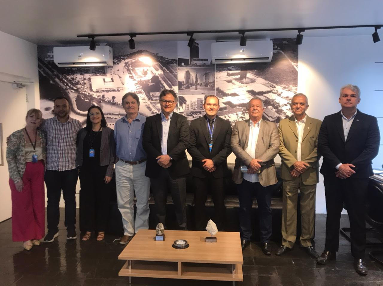 Um grupo de nove pessoas posando para uma foto, todas de pé, em frente a uma parede decorada com imagens em preto e branco de uma cidade ou local histórico. As pessoas estão alinhadas lado a lado, algumas com crachás, em um ambiente de escritório formal. Ao centro da imagem, há uma mesa pequena com dois troféus ou artefatos em exibição.