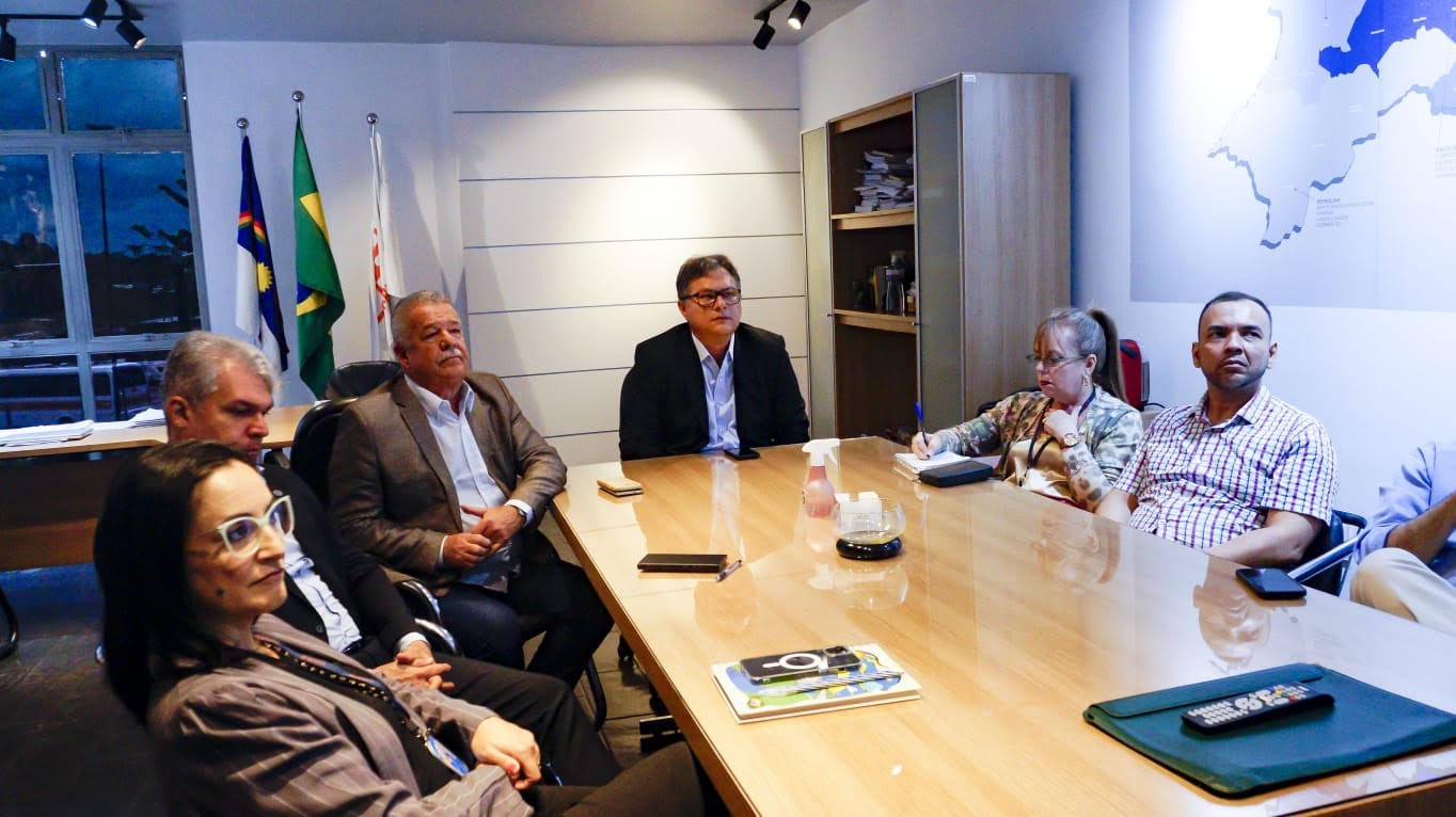 Um grupo de seis pessoas está sentado ao redor de uma mesa de reuniões de madeira, em um escritório. Ao fundo, há três bandeiras em suportes (provavelmente do Brasil, um estado e uma organização). As pessoas parecem atentas, possivelmente participando de uma reunião ou discussão. Entre os presentes, há uma mulher tomando notas. O ambiente tem iluminação embutida no teto e janelas à esquerda, com uma vista noturna.