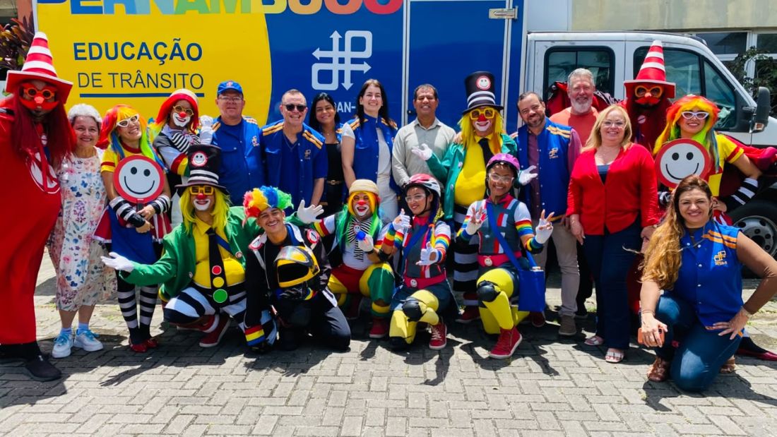 **Descrição de Acessibilidade (Texto Alternativo / Alt Text):** Fotografia em plano aberto mostrando um grupo de cerca de vinte pessoas posando juntas ao ar livre em frente a um veículo da Educação de Trânsito do DETRAN-PE. O grupo é composto por integrantes da equipe institucional com coletes azuis e por arte-educadores caracterizados como palhaços e personagens lúdicos, vestindo roupas bem coloridas, perucas, chapéus e maquiagens divertidas. Parte do grupo está em pé e outra parte agachada à frente. Ao fundo, vê-se um furgão branco com a inscrição "Educação de Trânsito".