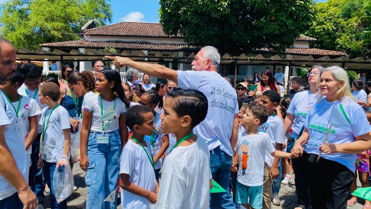 A imagem retrata um grupo de pessoas reunidas em frente a um prédio, com árvores visíveis ao fundo. * Uma multidão de pessoas: + A maioria dos indivíduos está vestindo camisetas brancas. + Algumas pessoas têm cordões verdes ao redor do pescoço. + Há uma pessoa que parece ser mais velha do que as outras. * Árvores: + Duas árvores grandes são visíveis atrás da multidão. + Elas parecem ser árvores decíduas com folhagem exuberante. * Prédios: + Ao fundo, há um prédio com um telhado de telhas. + Ele tem várias janelas ao longo de sua fachada. Em geral, a imagem sugere que o encontro pode estar relacionado a um evento ou atividade que ocorre no local, possivelmente envolvendo crianças, dado a presença de indivíduos mais jovens na multidão.