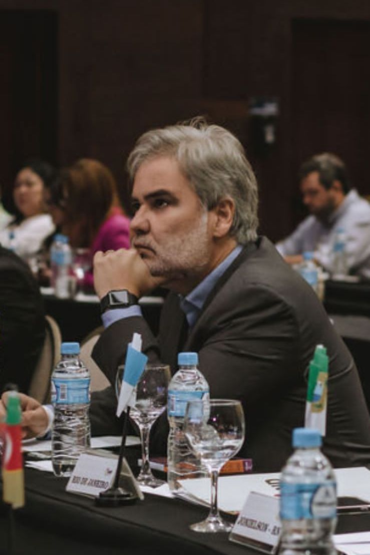 Na foto, o Diretor Presidente do DETRAN-PE, Gustavo Carneiro Leão, de perfil, com a mão esquerda no queixo e com teu cotovelo encostado na mesa. Na foto, ele está ao centro da imagem, sentado na cadeira. Essa imagem é do 74º Encontro Nacional dos Detrans, que aconteceu em Florianópolis (SC).