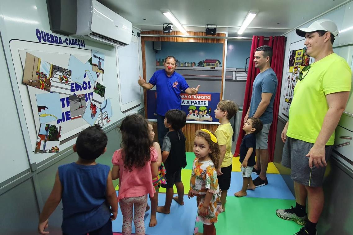 Na foto, um educador de trânsito mostra as crianças sobre atividade sobre educação de trânsito. Alguns pais observam ao fundo.