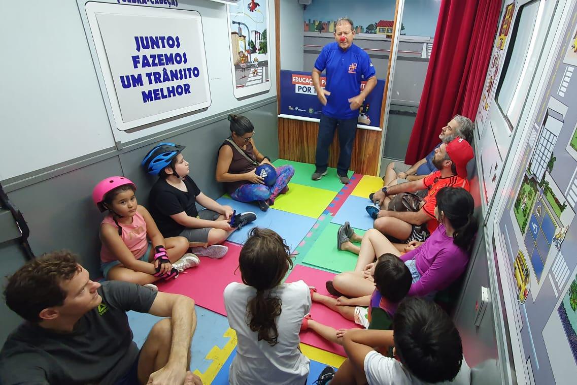 Na foto, educador de trânsito explicando para crianças e pais sobre as leis e o respeito no trânsito. As crianças e pais estão sentado em forma de círculo, enquanto o educado está em pé.