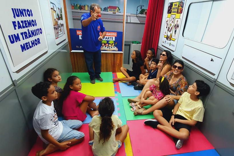 Na foto, educador de trânsito explicando para crianças e pais sobre as leis e o respeito no trânsito. As crianças e pais estão sentado em forma de círculo, enquanto o educado está em pé.