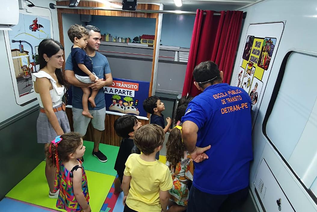 Na foto, um educador de trânsito mostra as crianças sobre atividade sobre educação de trânsito. Alguns pais observam ao fundo.