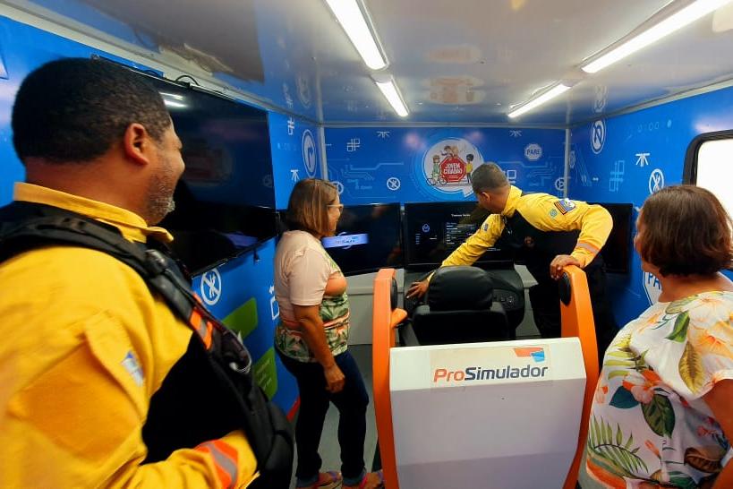 Na foto, dois agentes de trânsito do DETRAN-PE apresentam o simulador de trânsito para duas professoras.