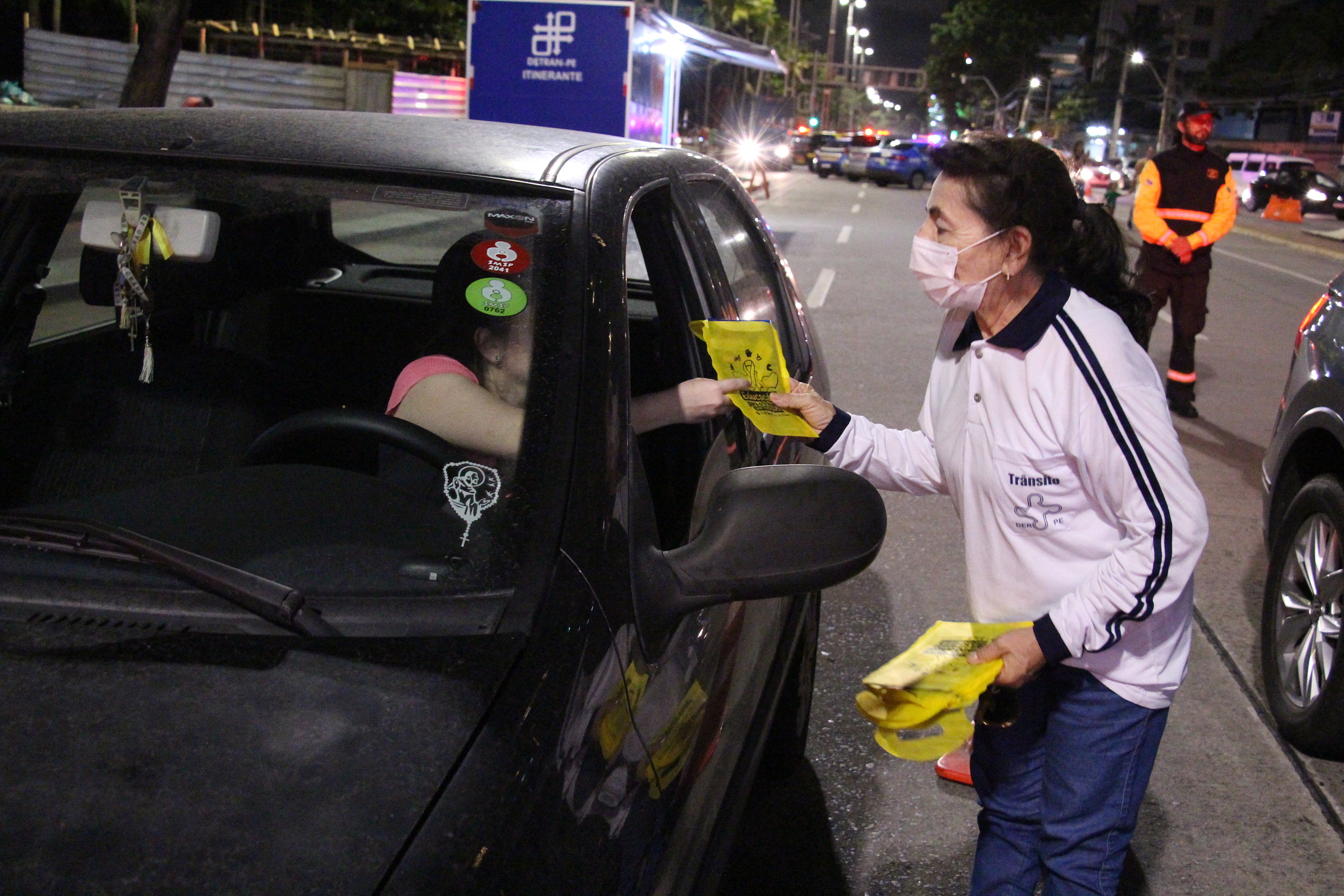 Na foto, agente feminina do DER entregando brinde com panfletos sobre educação no trânsito para motorista. 