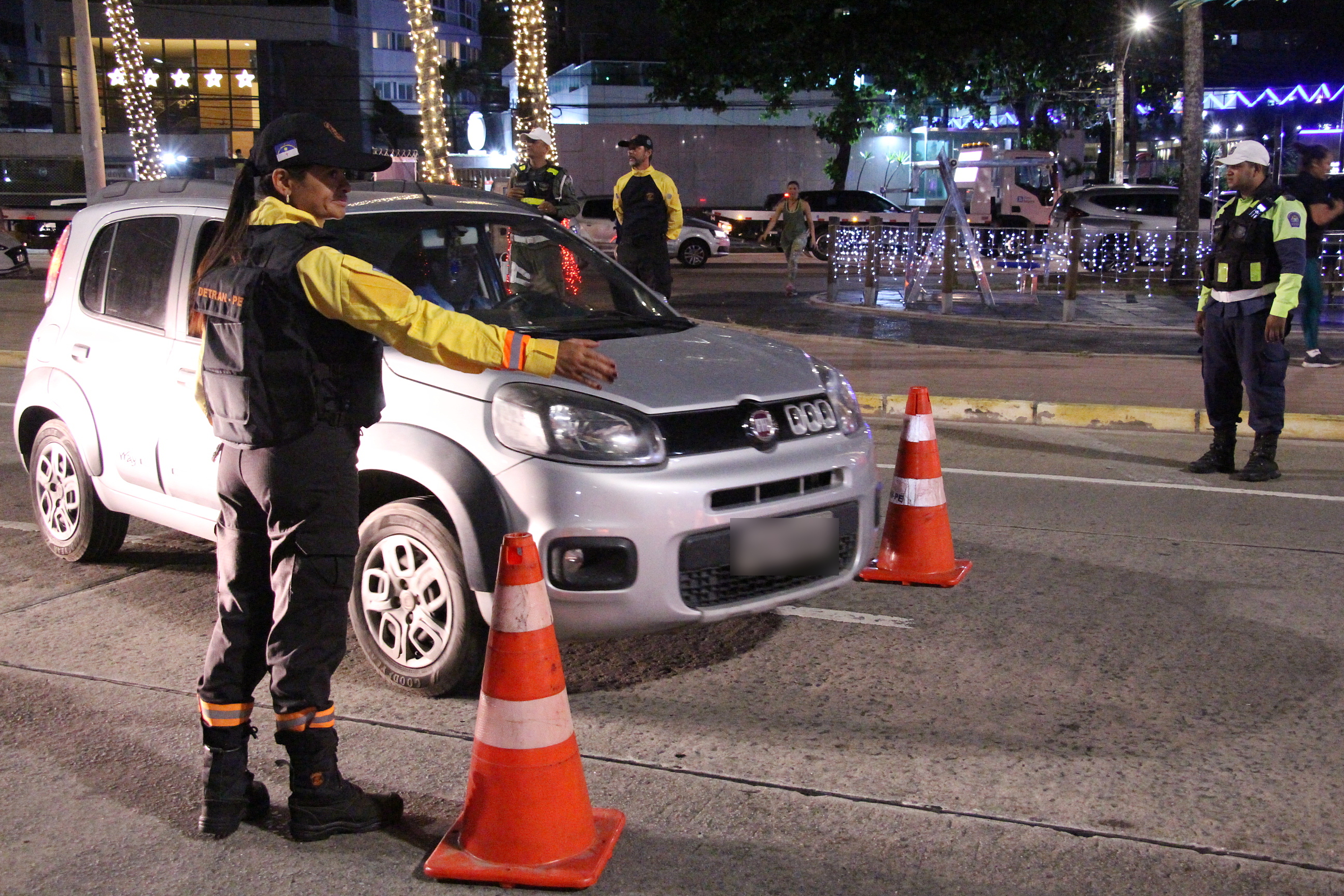 Na foto, agente de trânsito feminina do DETRAN-PE, sinalizando para veículo estacionar em local direcionado. Ao fundo, agentes de trânsito da CTTU e DETRAN-PE.