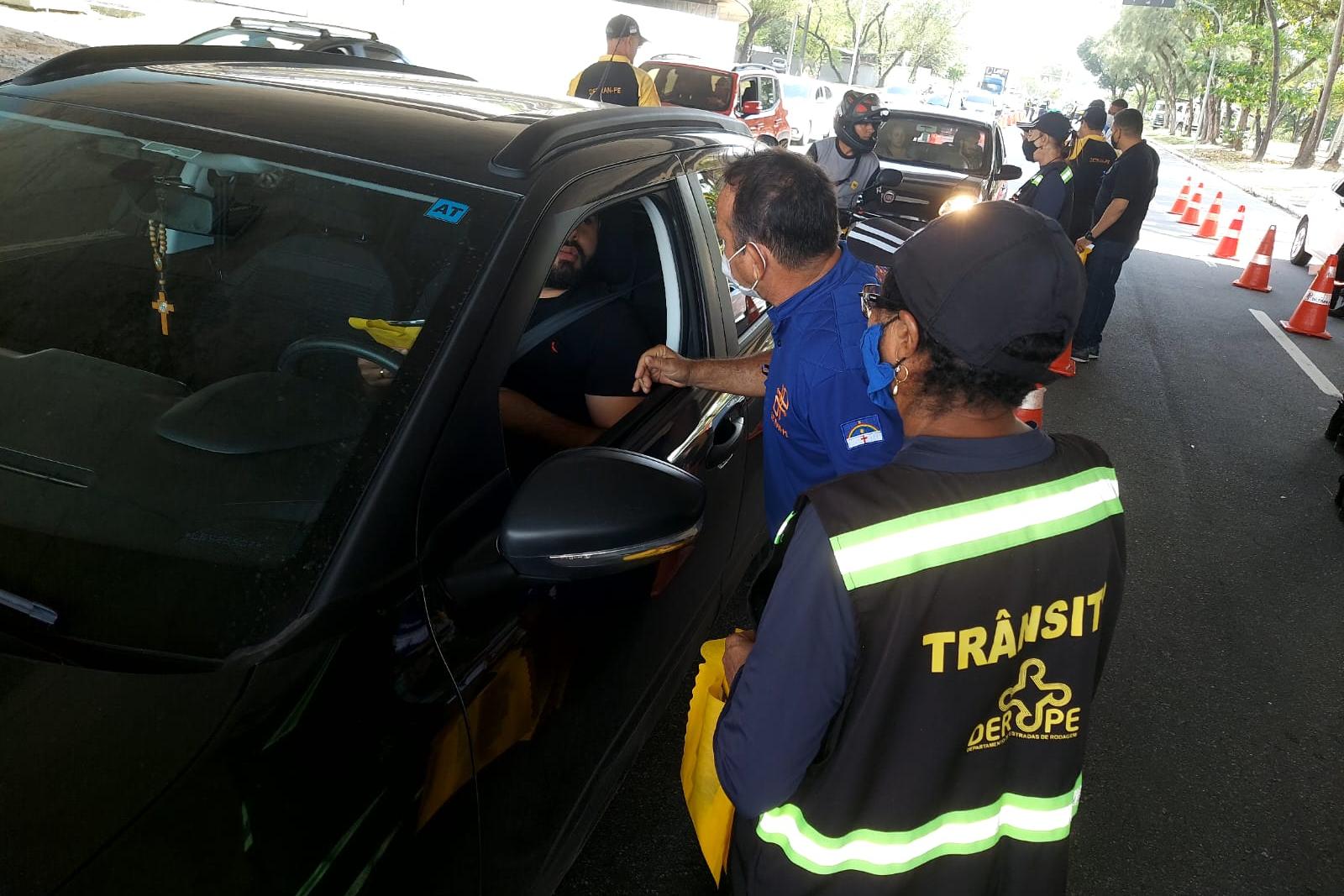 Na foto, educador de trânsito do DETRAN-PE e agente do DER abordando motorista em ação educativa. Ao fundo, agente de trânsito do DETRAN-PE e um outro carro sendo abordado na ação educativa. 