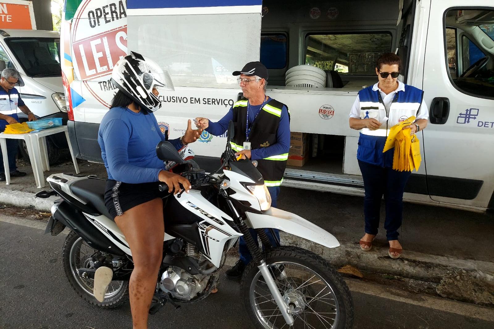 Na foto, agente do DER abordando motociclista em ação educativa e, ao seu lado, agente da Operação Lei Seca – OLS. Ao fundo o caminhão da OLS.