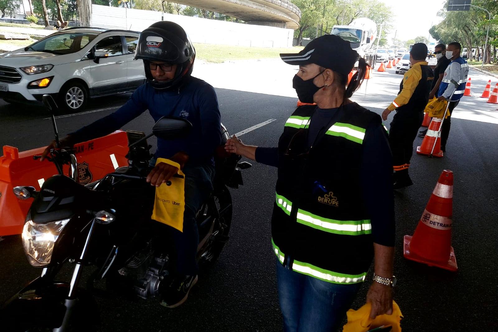 Na foto, agente do DER abordado motociclista em ação educativa. Ao fundo, agente de trânsito do DETRAN-PE e educadores da OLS.
