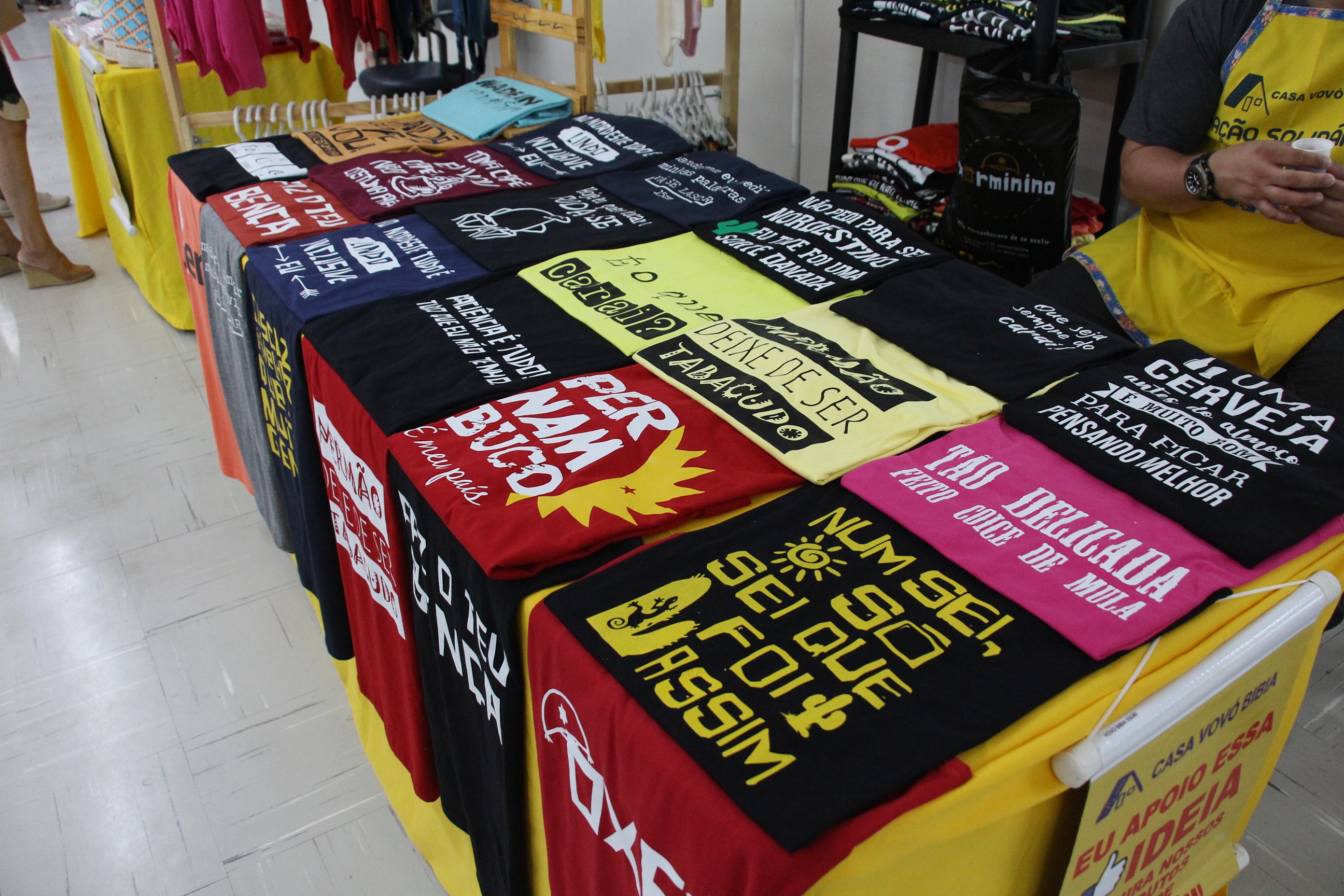 Na foto, uma barraca com exposição de camisas personalizadas, de várias cores, com ditados nordestinos.