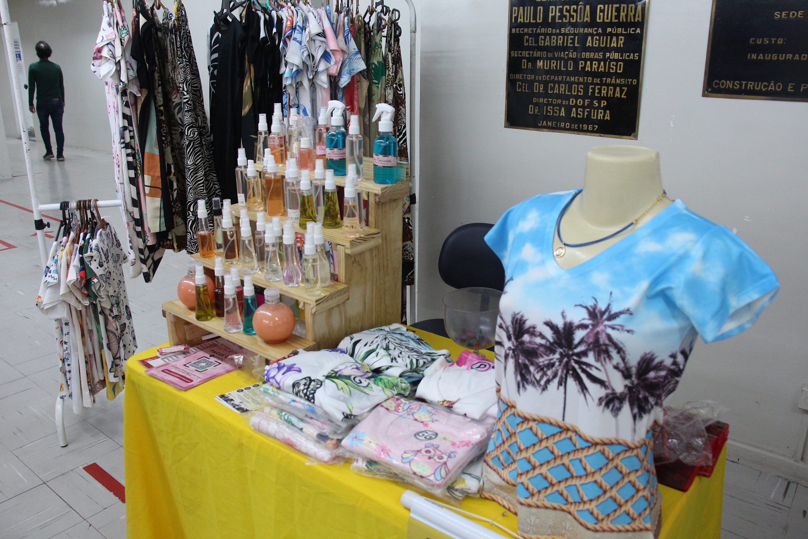 Na foto, uma barraca com exposição de camisa e perfumaria. Ao fundo, uma arara de roupa com vestidos expostos.