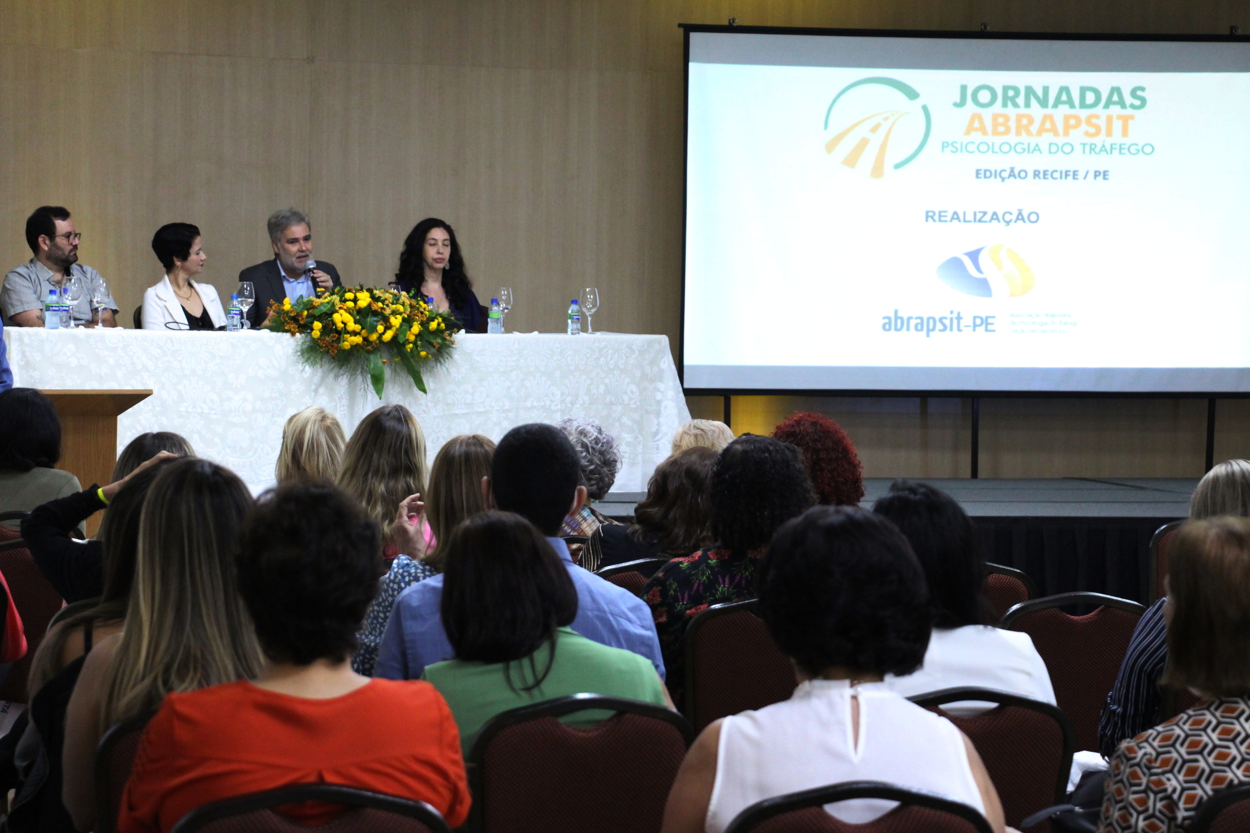 Na foto, o  Diretor Presidente do DETRAN-PE, Gustavo Carneiro Leão, ao lado da Gerente de Psicomédica do DETRAN-PE, Juliana Guimarães; do Conselho Regional de Psicologia-PE, Marcos Murcabel; e da chefe da Unidade de Psicologia do Detran-PE, Sheila Azevedo, discursando na abertura da II Jornada Pernambucana de Psicologia de Tráfego da Abrapsit, no Beach Class Convention, em Boa Viagem. Em segundo plano, a plateia.  Na lateral da foto, um telão com a frase “Jornadas Abrapsit, psicologia do tráfego”, abaixo, a logo da Abrapsit.
