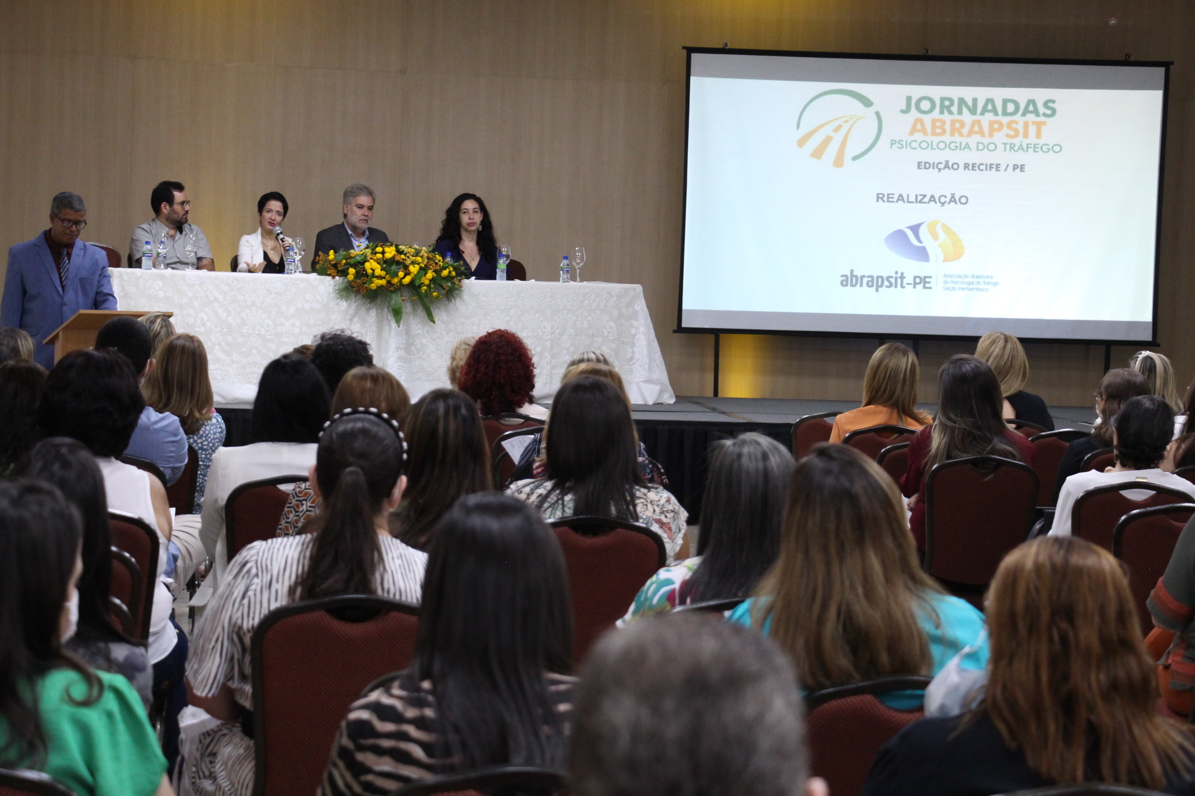 Na foto, a Gerente de Psicomédica do DETRAN-PE, Juliana Guimarães, ao lado do Diretor Presidente do DETRAN-PE, Gustavo Carneiro Leão; do Conselho Regional de Psicologia-PE, Marcos Murcabel; e da chefe da Unidade de Psicologia do Detran-PE, Sheila Azevedo, discursando na abertura da II Jornada Pernambucana de Psicologia de Tráfego da Abrapsit, no Beach Class Convention, em Boa Viagem. Em segundo plano, a plateia.  Na lateral da foto, um telão com a frase “Jornadas Abrapsit, psicologia do tráfego”, abaixo, a logo da Abrapsit.