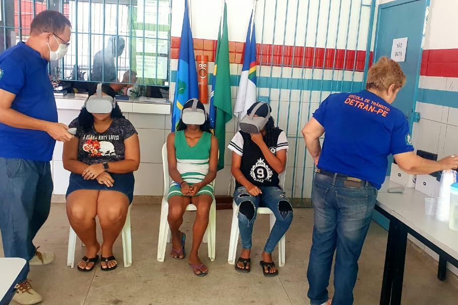 Na foto, educadores de trânsito do DETRAN-PE auxiliando três alunos da Escola Municipal Manoel da Rosa, em Paudalho, quando estão fazendo o uso do óculos de realidade virtual, acessório que leva o usuário para uma viagem virtual, partindo de um bar, após ingerir bebida alcoólica, e segue por ruas e avenidas, finalizando de maneira trágica.