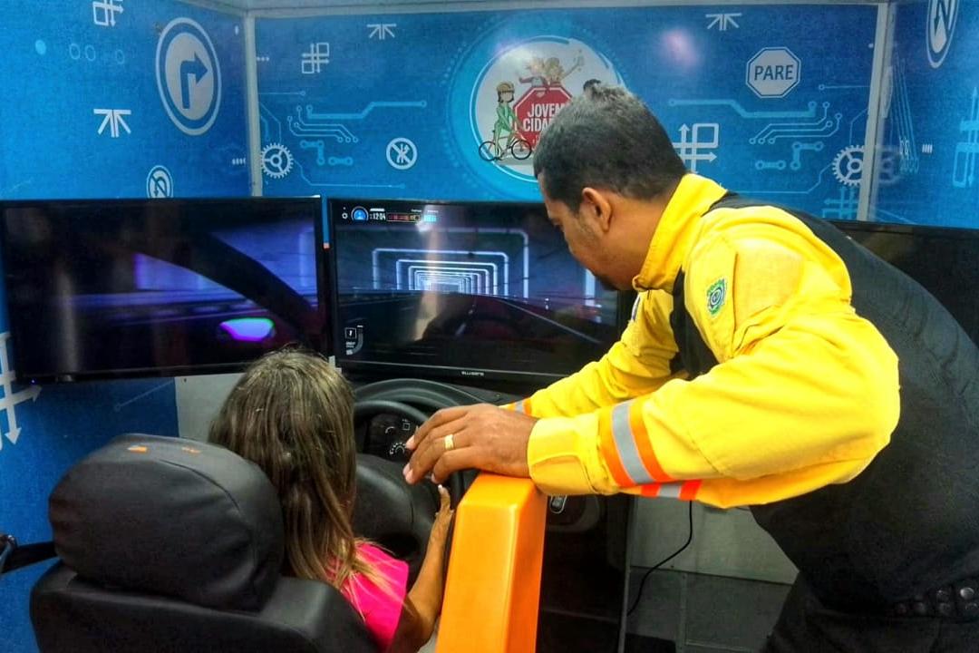 Na foto, agente de trânsito do DETRAN-PE auxiliando uma aluna da Escola Municipal Manoel da Rosa, em Paudalho, no simulador de direção, que apresenta uma visão aproximada das condições de dirigir estando sob efeito de álcool. O equipamento, composto por volante, pedais, alavanca de marcha e monitores que mostram a estrada, representa sensações como distorções na via, lentidão de reflexos, visão embaçada e velocidade fora dos limites permitidos.