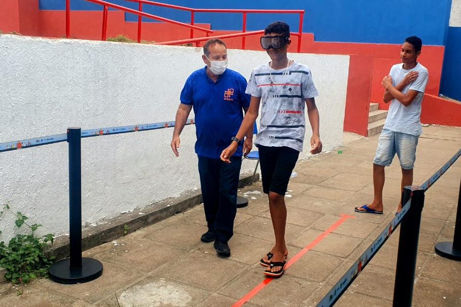 Na foto, educador de trânsito do DETRAN-PE auxiliando um aluno da Escola Municipal Manoel da Rosa, em Paudalho, que está fazendo o uso do óculos que simulam embriaguez. A peça transmite a noção de tontura, vista turva e desequilíbrio, condições habituais após o uso de álcool.