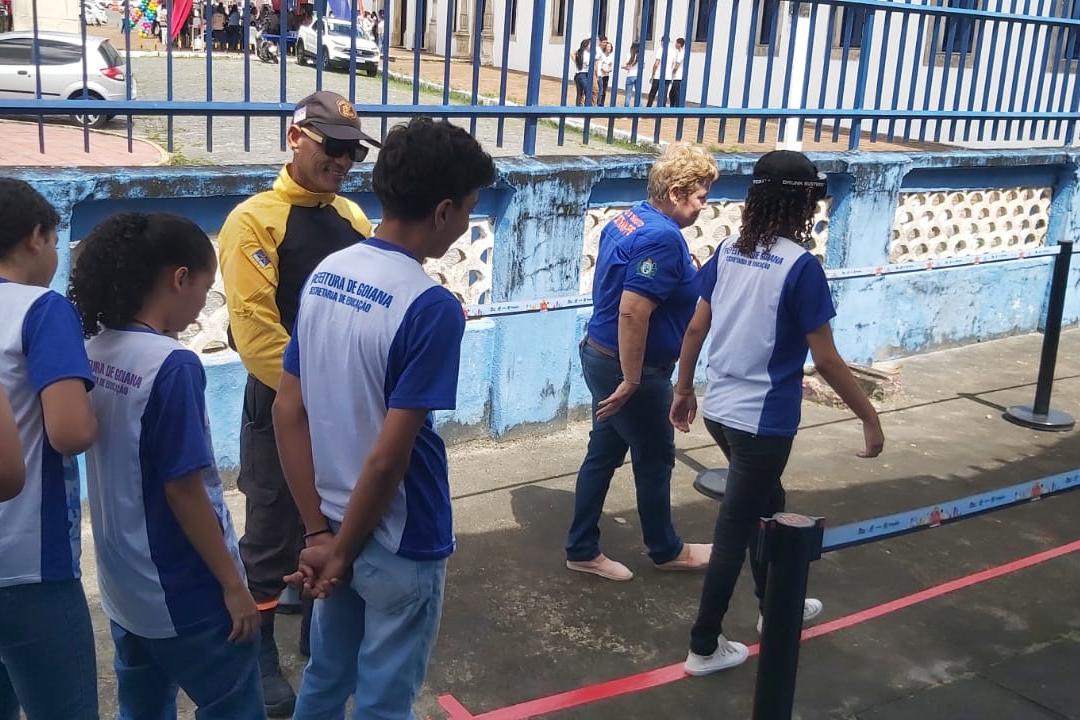 um aluno fazendo o uso do óculos que simula o estado de embriaguez, outros alunos ao redor, um agente de trânsito e um educador de trânsito do DETRAN-PE auxiliando na ação. 