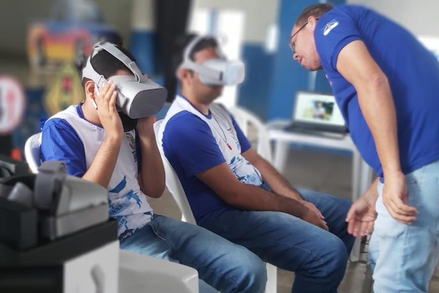 dois alunos fazendo o uso do óculos de realidade virtual, ao lado deles, ajudando, está um educador de trânsito