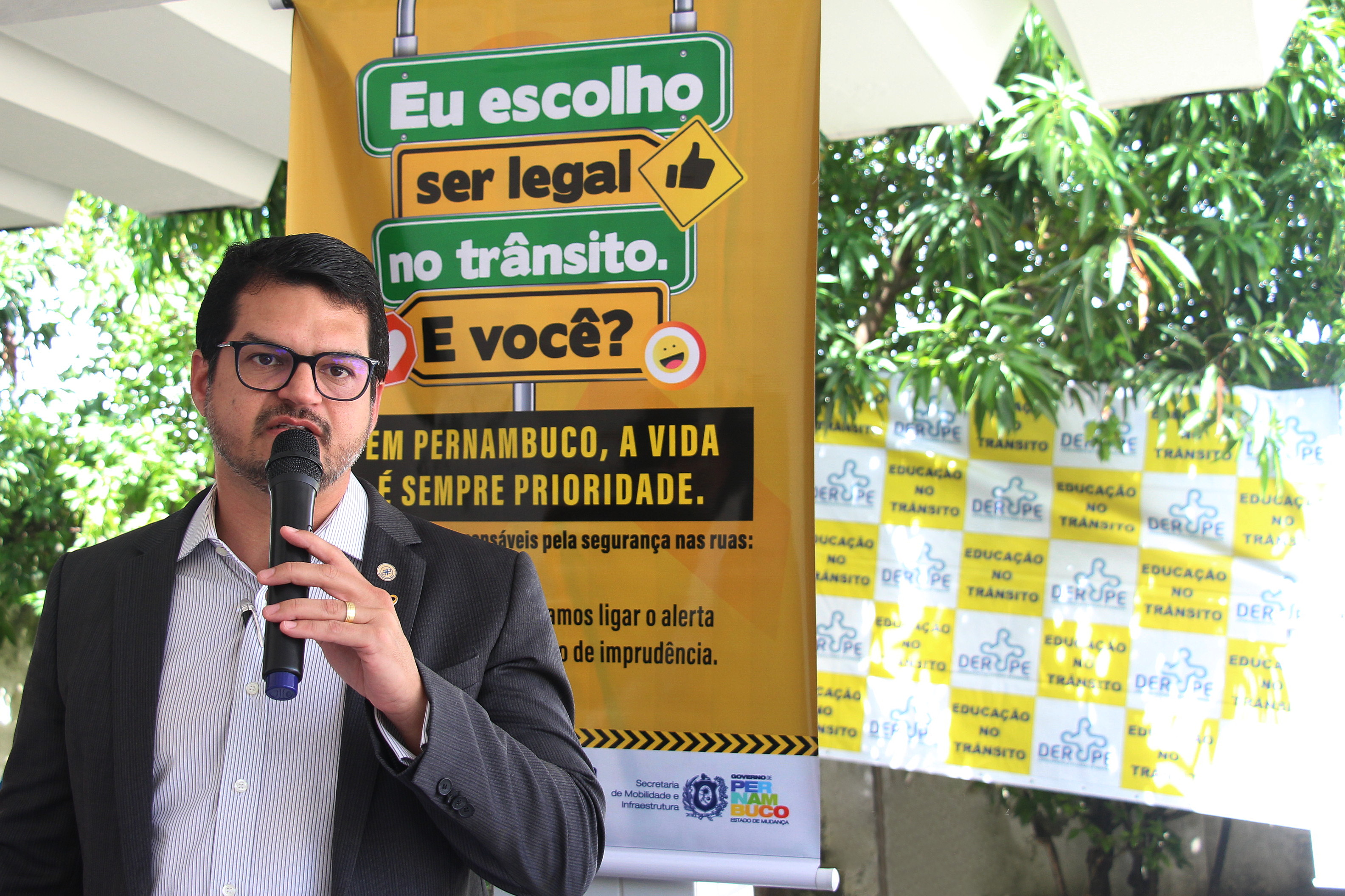 Na foto, o Diretor Presidente do DETRAN-PE, Carlos Fernando Ferreira, realizando sua fala na abertura do Movimento Maio Amarelo na sede do DER. Ao fundo, banner do Maio Amarelo com a frase: “Eu escolho ser legal do trânsito. E você?”