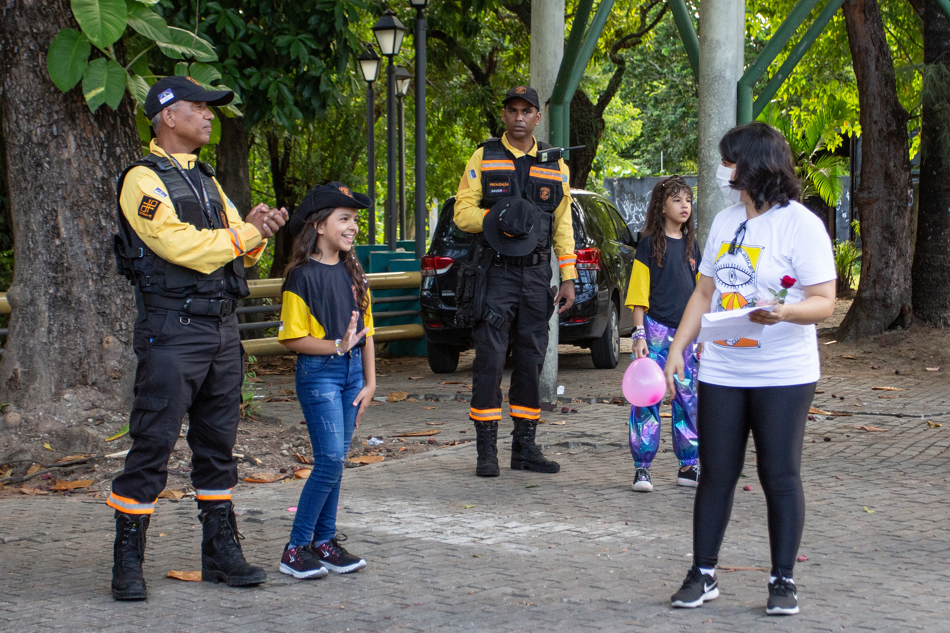 Na foto, dois agentes de trânsito fardados com duas meninas usando a farda de agentes de trânsito do DETRAN-PE. Uma das meninas está acenando para uma mulher. A ação aconteceu em frente ao Parque da Jaqueira e foi em homenagem ao Dia das Mães.