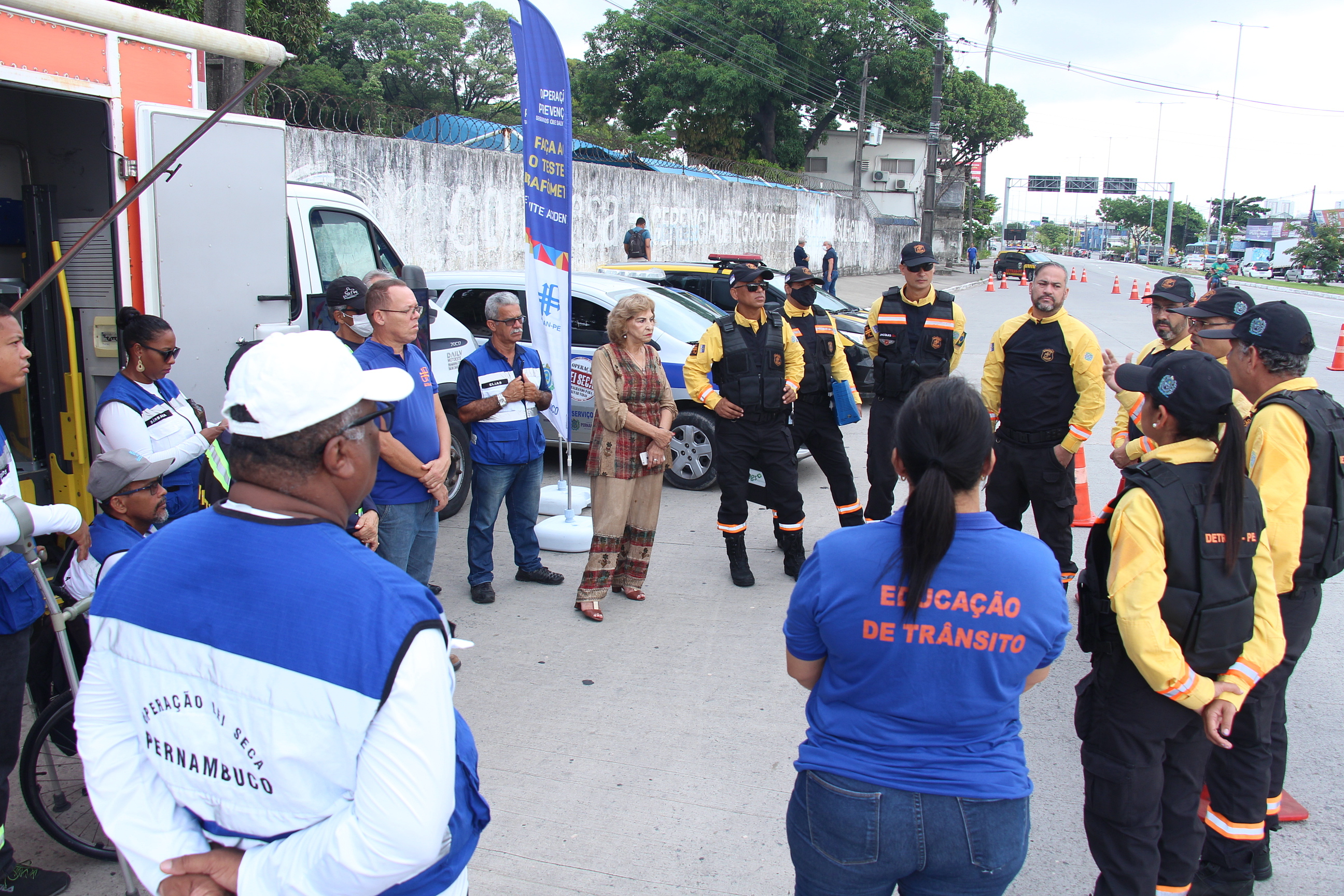 Na foto, educadores e agentes de trânsito do DETRAN-PE, equipe da Operação Lei Seca e do DER, reunidos em círculo, recebendo orientações sobre os procedimentos a serem realizados na blitz. Ao fundo, veículos da Operação Lei Seca e da fiscalização de trânsito do DETRAN-PE. 