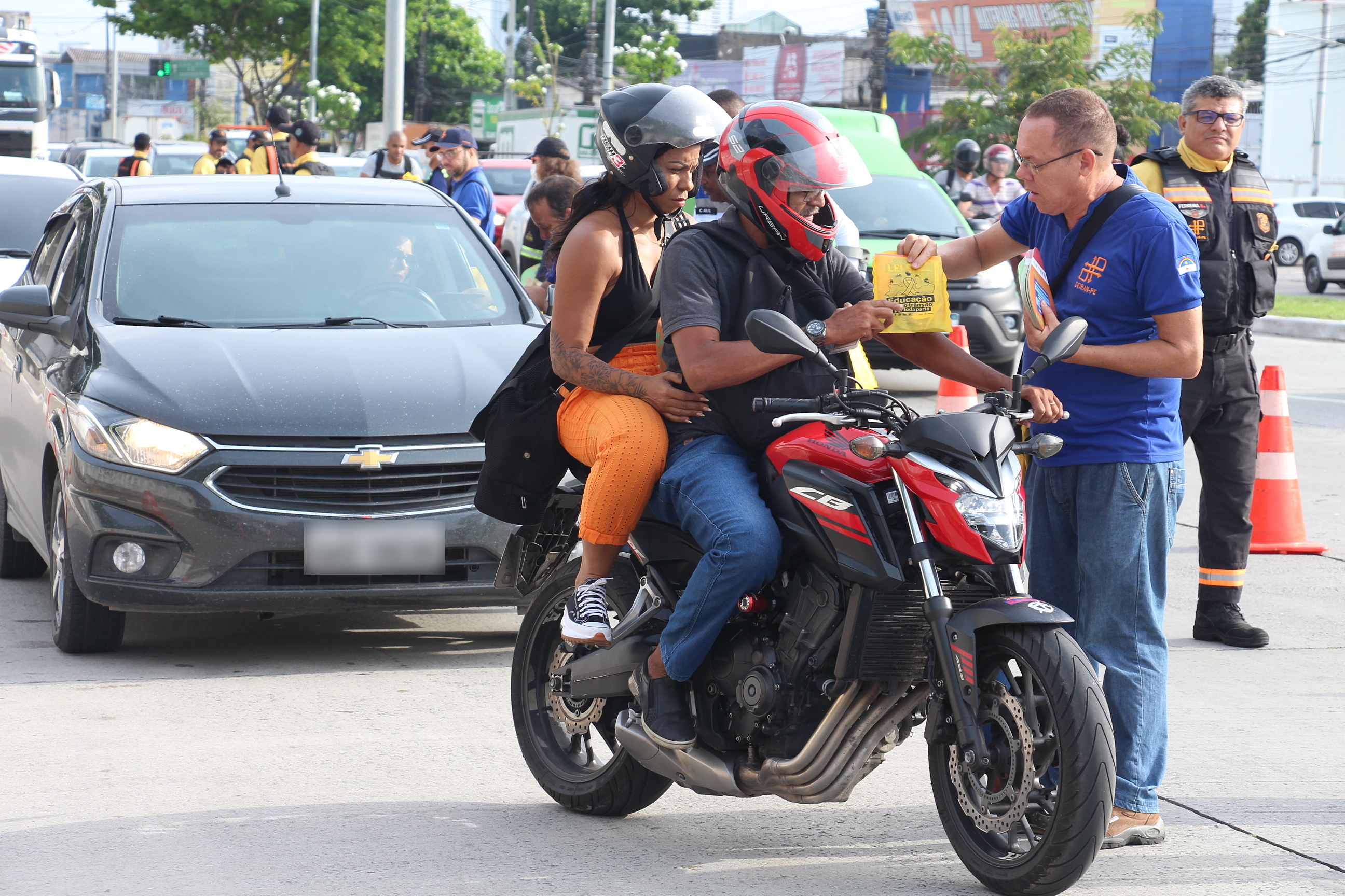 Na foto, um motociclista e sua carona sendo abordados por educador de trânsito, quando ele entregou material educativo.