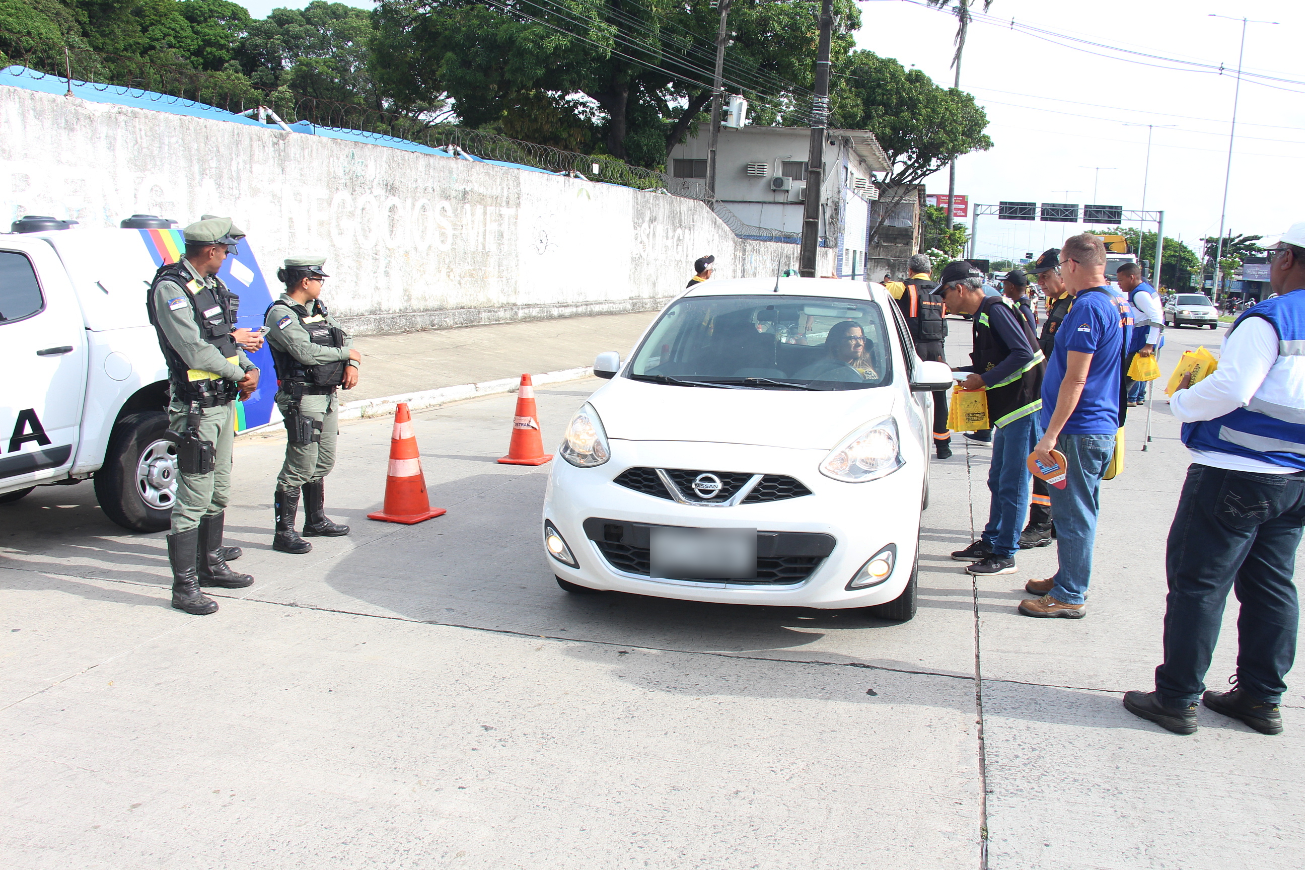 Na foto, um carro branco sendo abordado, quando agente do DER e educador de trânsito do DETRAN-PE alertam para os cuidados do trânsito. Também estão na foto, policiais do BPTran. 