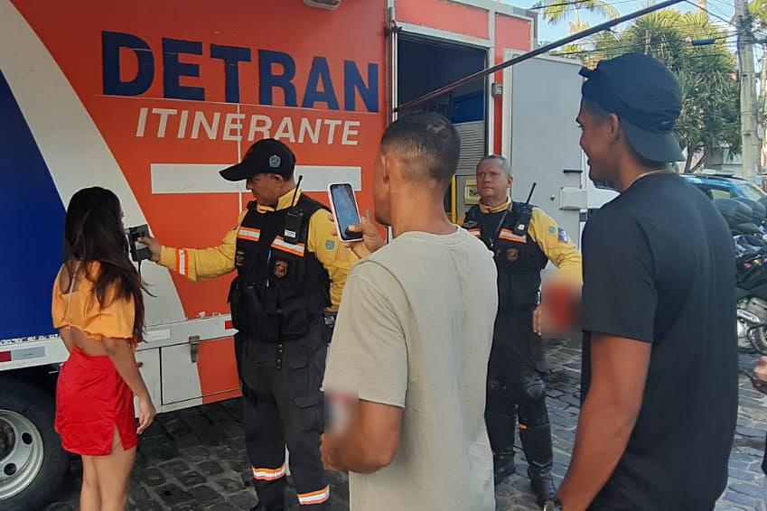 Agente de Trânsito realizando bafómetro em mulher que estava participando das prévias carnavalescas de Olinda. Ao fundo, o caminhão do DETRAN Itinerante. Dois homens estão filmando a ação. 