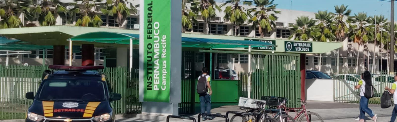 Na foto, veículo da fiscalização de trânsito do DETRAN-PE em frente ao IFPE. Na frente, estão estacionadas bicicletas. Alunos estão entrando no colégio. 