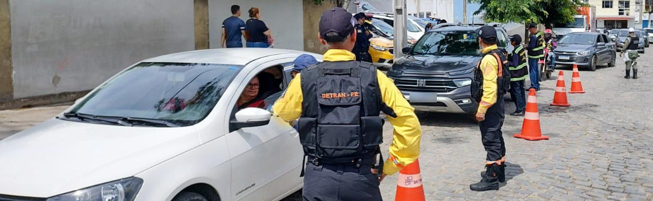 Na foto, três veículos abordados, um na cor branco e dois na cor cinza escuro. Dois agentes de trânsito do DETRAN-PE estão em primeiro plano na foto. Ao fundo, dois servidores do DER estão abordando o veículo de cor cinza escuro. Mas ao fundo da foto, um policial militar está organizando o trânsito.