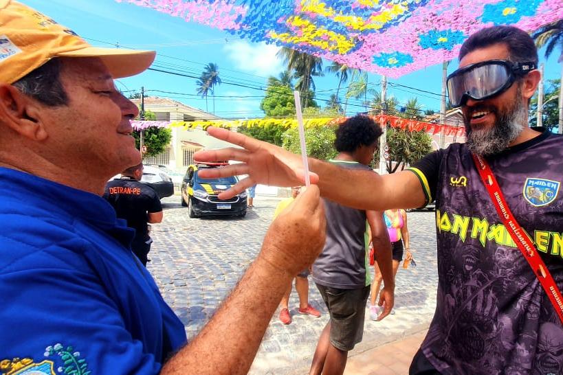Na foto, educador de trânsito do DETRAN-PE realizando ação educativa com folião. Ele faz o uso do óculos que deixa com a sensação de embriaguez.