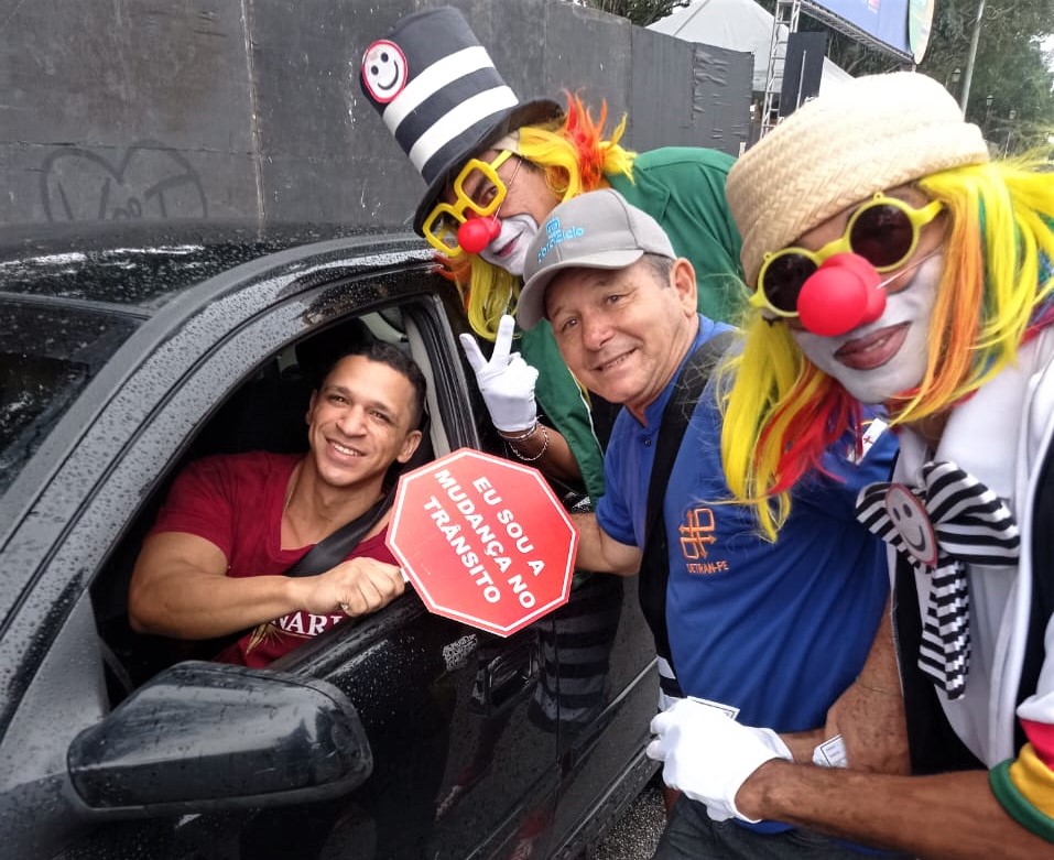 Fotografia colorida de motorista recebendo placa de educador do Detran-PE e palhaços da turma do Fom-Fom com a frase 