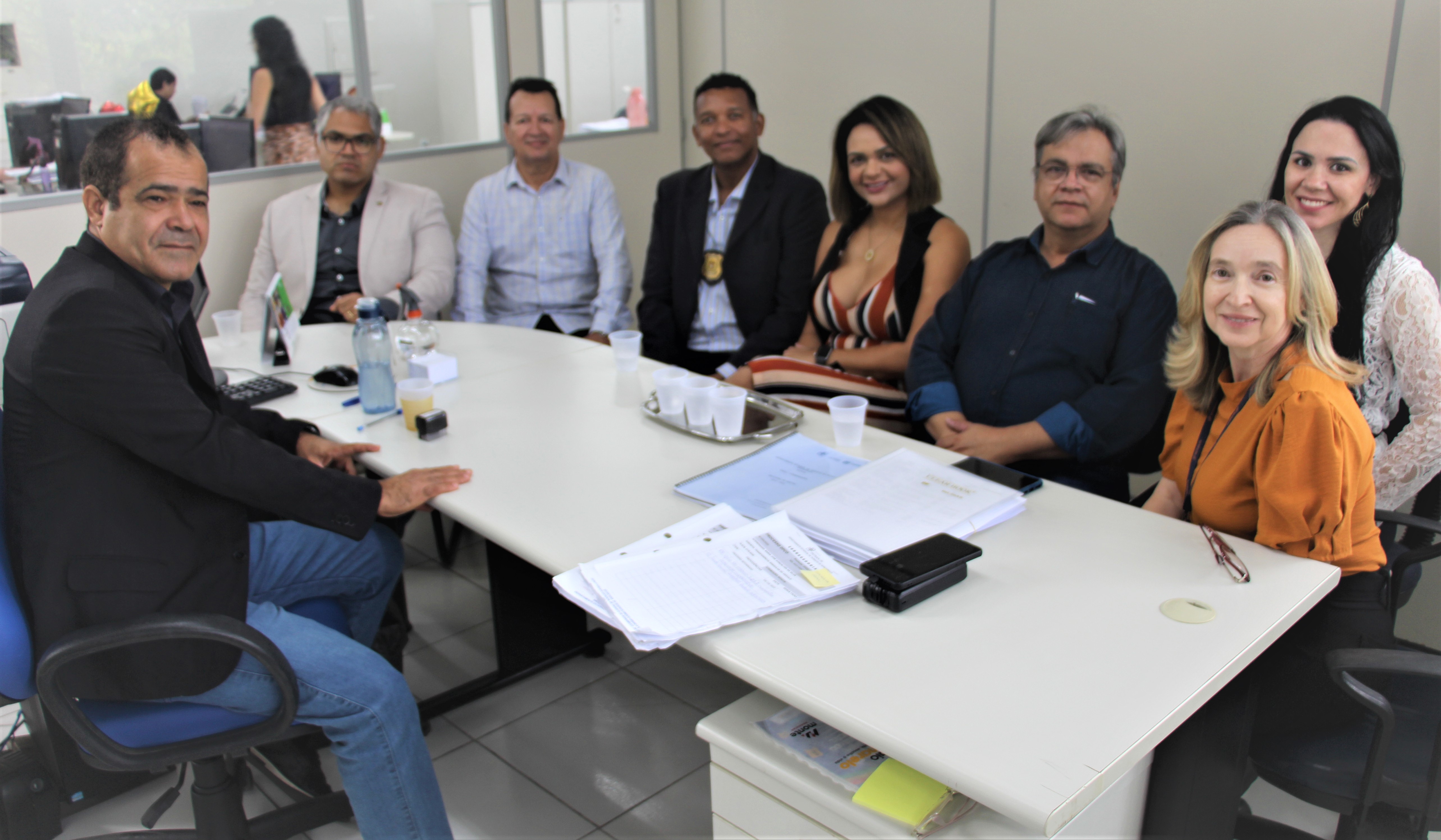 Fotografia colorida com participantes dispostos em volta de mesa de trabalho